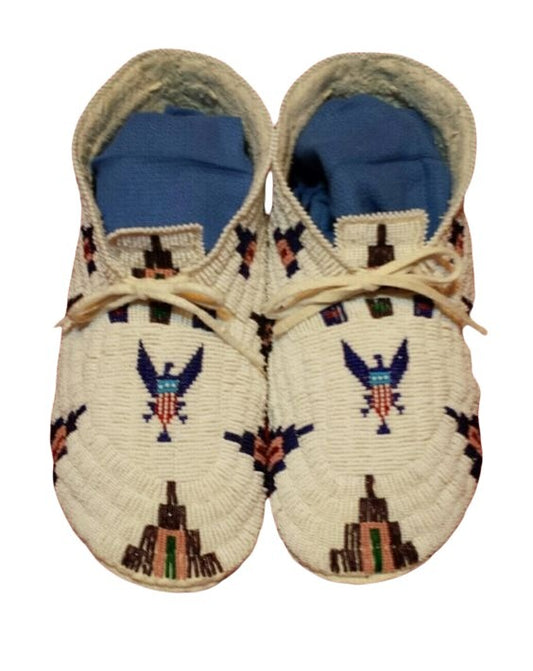 A pair of beaded moccasins featuring a design with eagles and geometric patterns, in a white, blue, and multicolor palette.
