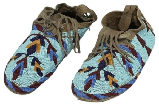 A pair of handmade beaded moccasins with a geometric pattern featuring blue, brown, and beige colors, with hand-stitched leather sole.