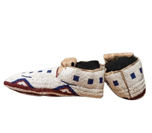 A pair of handmade beaded moccasins with a mix of brown, white, and red geometric patterned beads, featuring hand stitching and designed with a Native American style.
